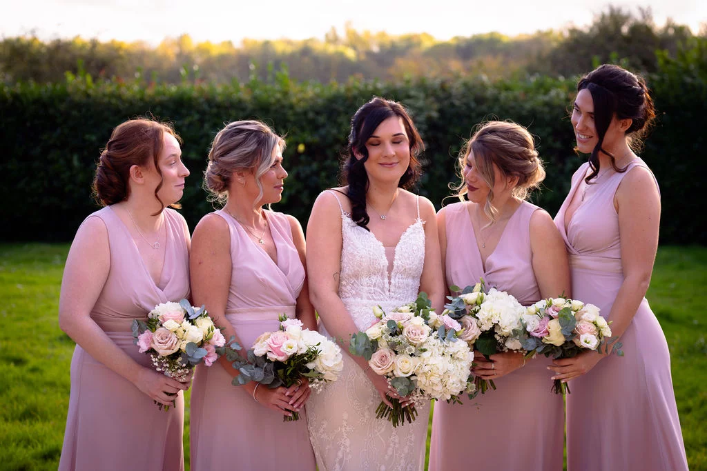 A radiant bride stands outdoors in a lace wedding gown holding a large white and pink bouquet, surrounded by her four bridesmaids wearing matching pink V-neck dresses during the golden hour.