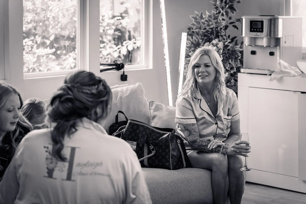 A mature woman sits on a sofa holding a glass of champagne during bridal morning preparations, while another person in a personalised robe is seen from behind.