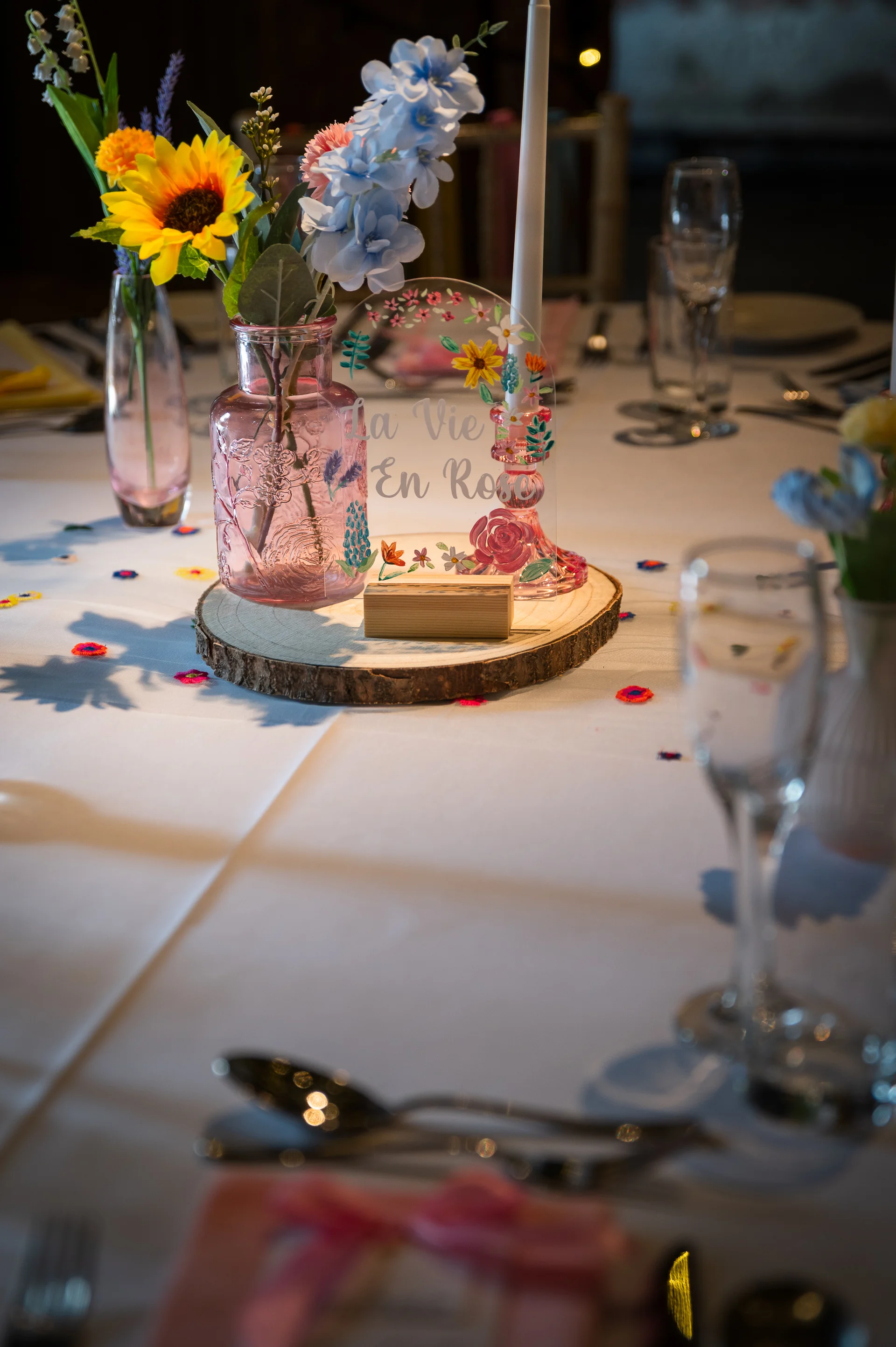 A charming wedding reception tablescape featuring a wooden slice centrepiece with a pink glass bud vase, colourful wildflowers, a taper candle, and an acrylic table sign.