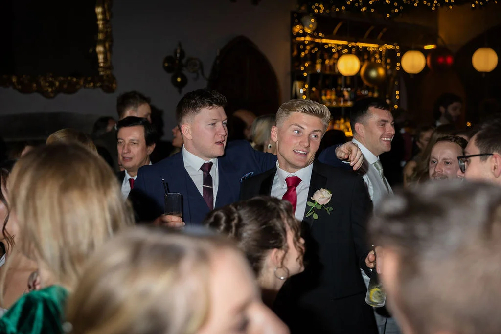 Groomsmen and wedding guests enjoy drinks and conversation during a lively evening reception in a warmly lit bar venue.