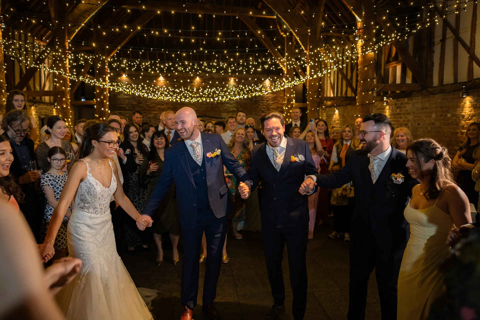 A smiling bride and a group of groomsmen hold hands in a circle on the dance floor inside a rustic barn illuminated by a stunning fairy light canopy.