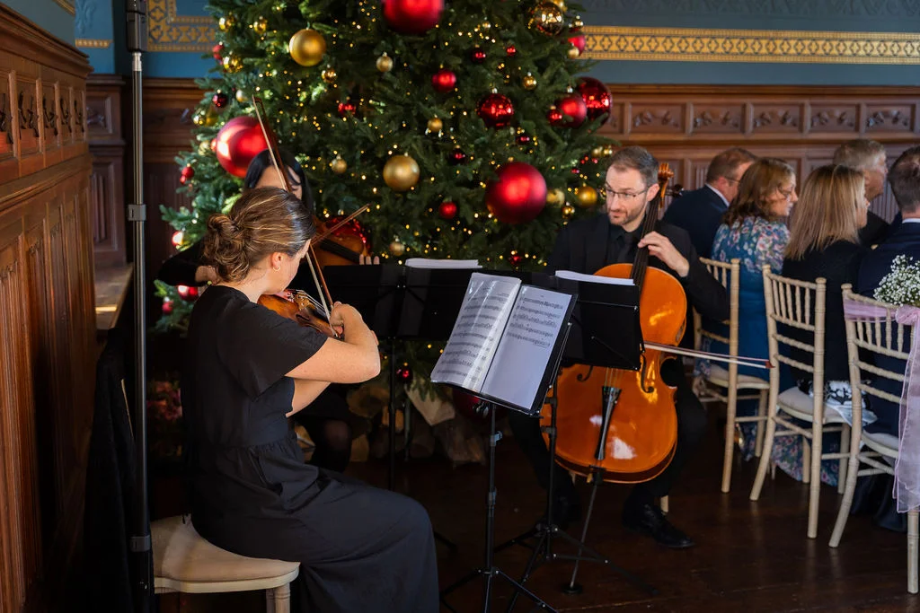 A live string trio performs background music in a panelled room decorated with a large Christmas tree and red and gold baubles during a festive wedding celebration.