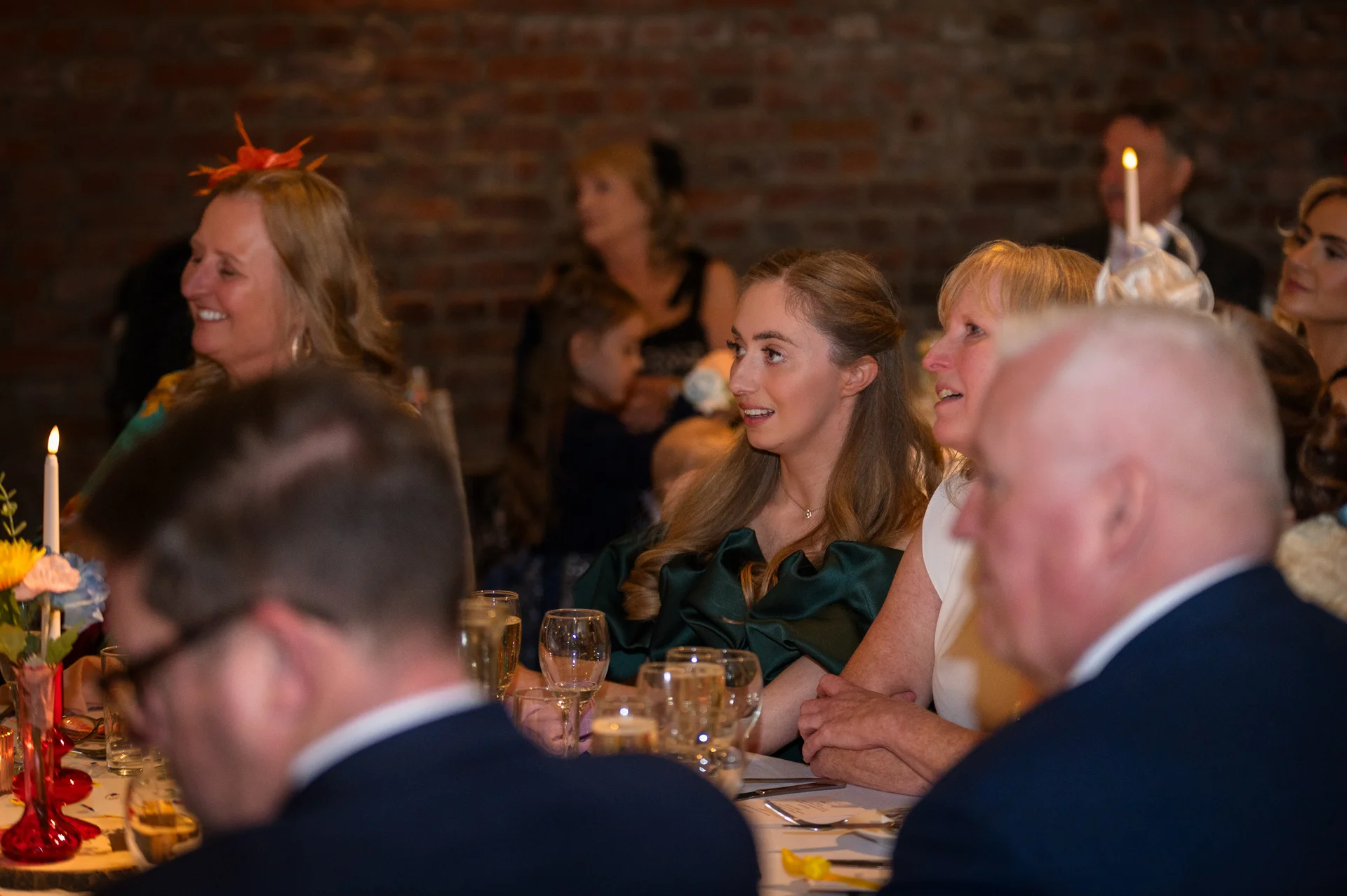 Wedding guests seated at tables inside a rustic brick venue, listening attentively and smiling during the wedding speeches.