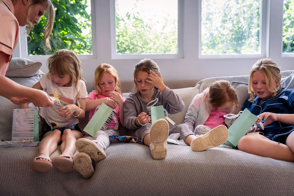 Several young girls sit on a large sofa opening small gift bags while getting ready in a bright, light-filled room.