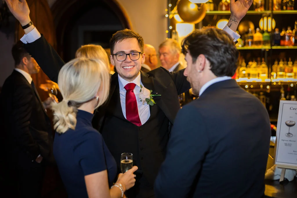 A smiling groom raises his arms in celebration while celebrating with wedding guests at a lively evening reception near a lit bar.