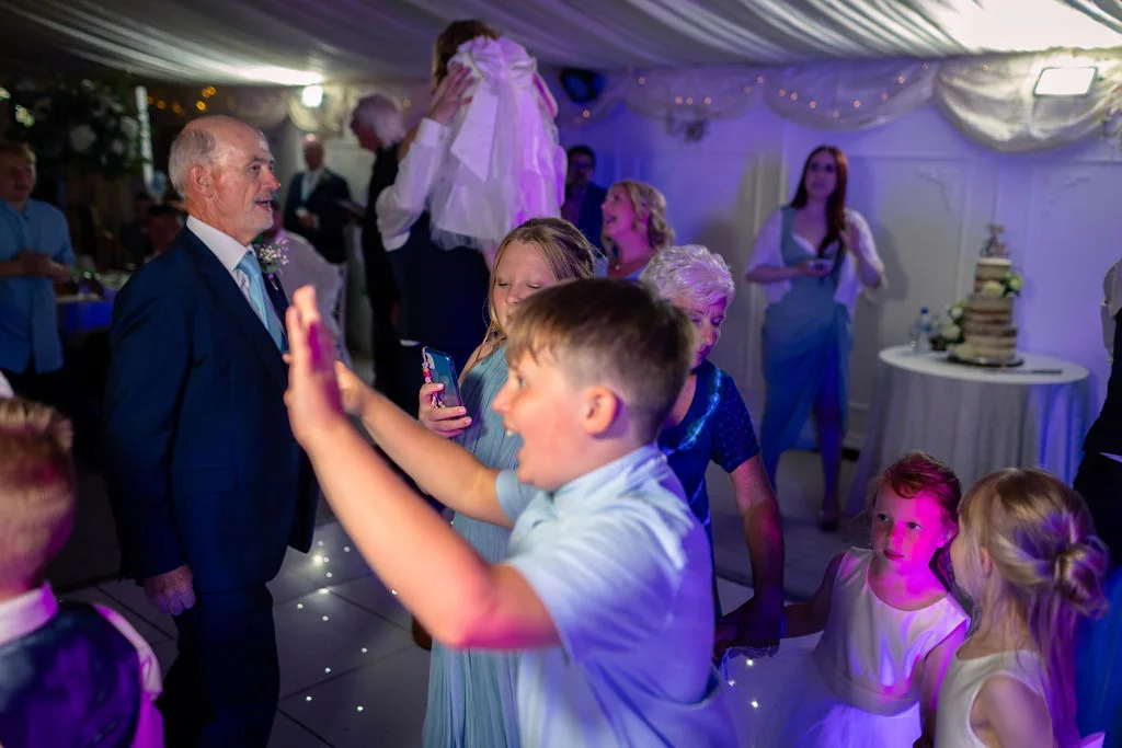 A joyful young boy dances with his hands in the air on an illuminated dance floor during a lively evening wedding reception.