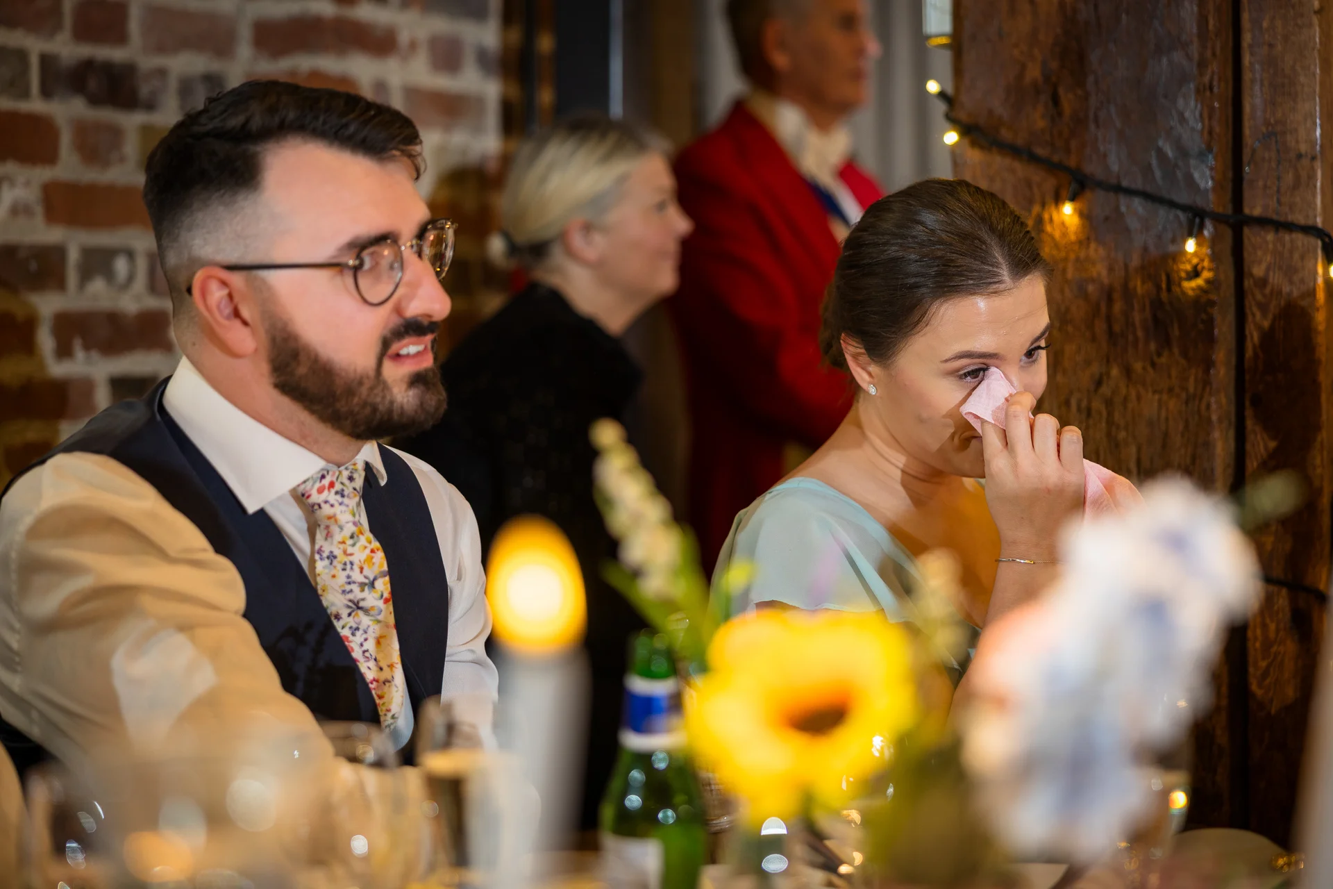 A guest is visibly moved, wiping away happy tears with a napkin during the wedding speeches at a rustic venue with exposed brick.