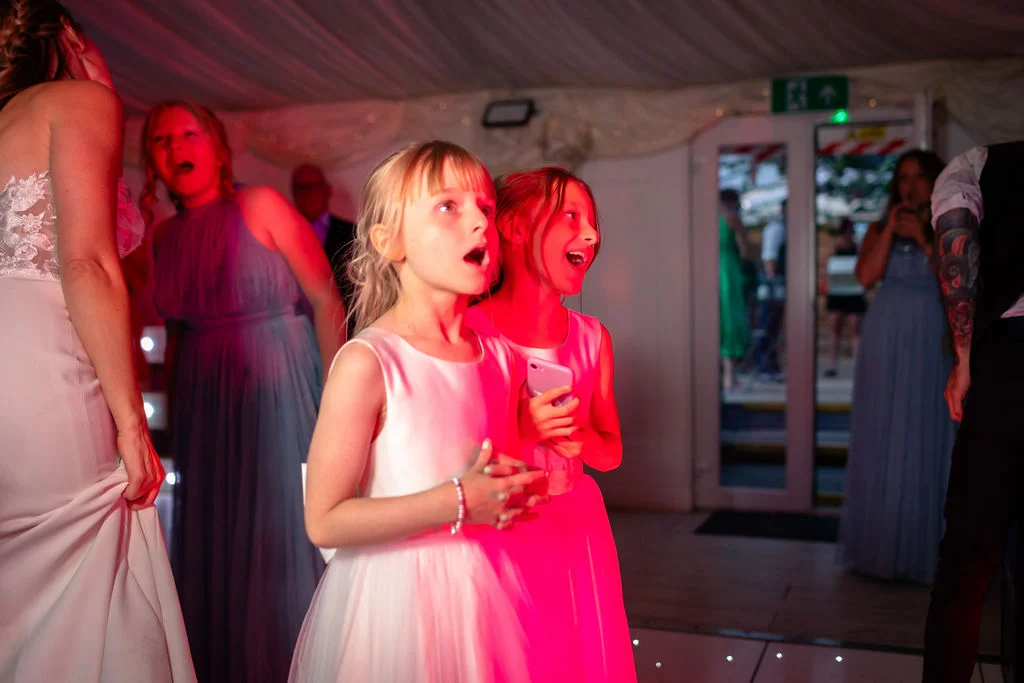 Two young flower girls in white dresses watch excitedly with open mouths under colourful disco lights at an evening wedding reception.