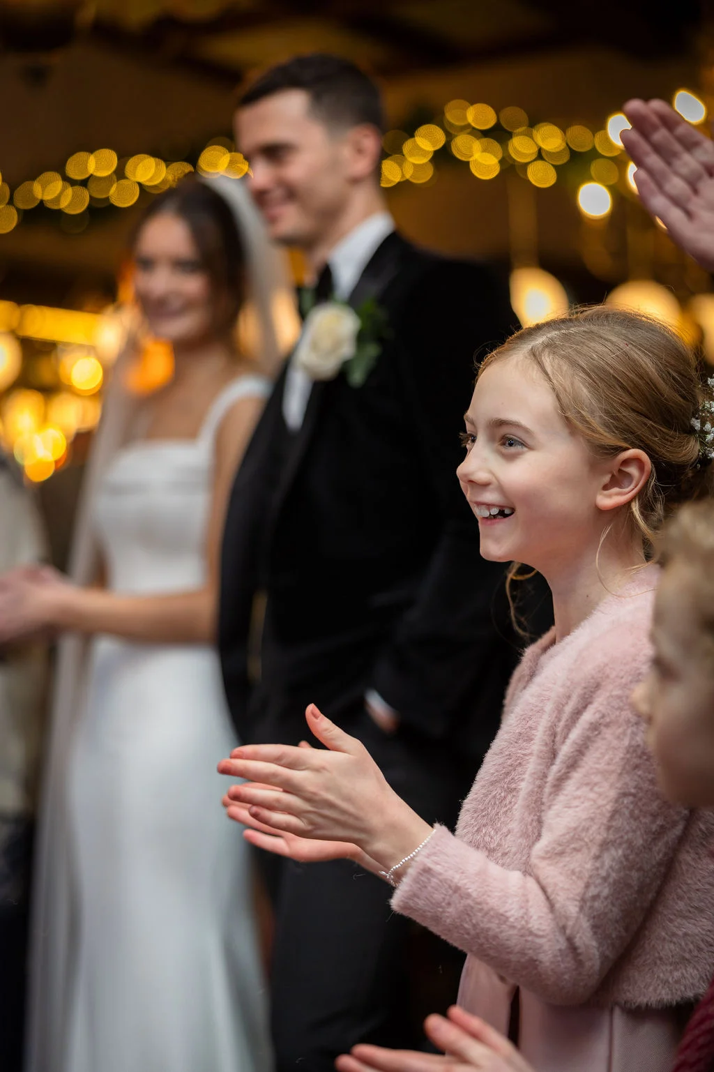 A young flower girl smiling and clapping enthusiastically while guests cheer, with the bride and groom blurred in the background.