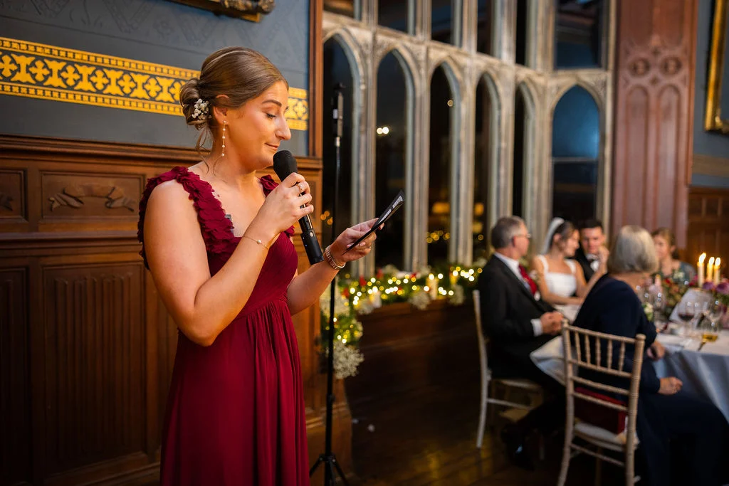 A bride gives a speech holding a microphone and notes in a grand wood-panelled hall during the wedding reception.