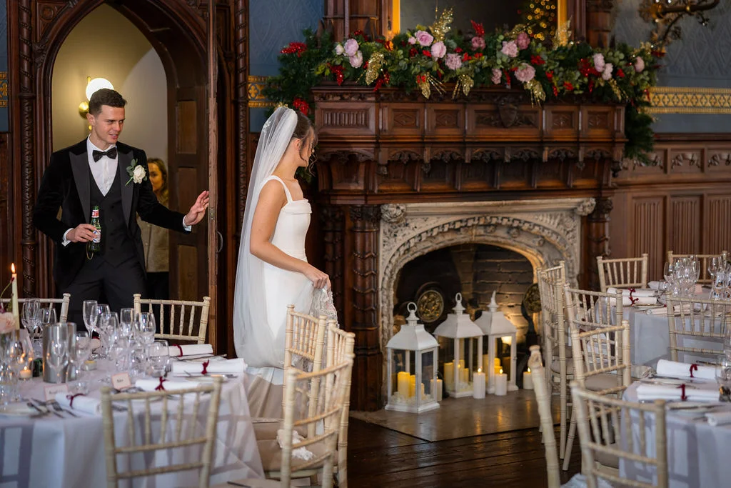 A groom in a black tuxedo holding a drink opens a wooden door as the bride in a sleek white gown with a chapel veil enters a grand room featuring a festive fireplace and elegantly set dining tables.