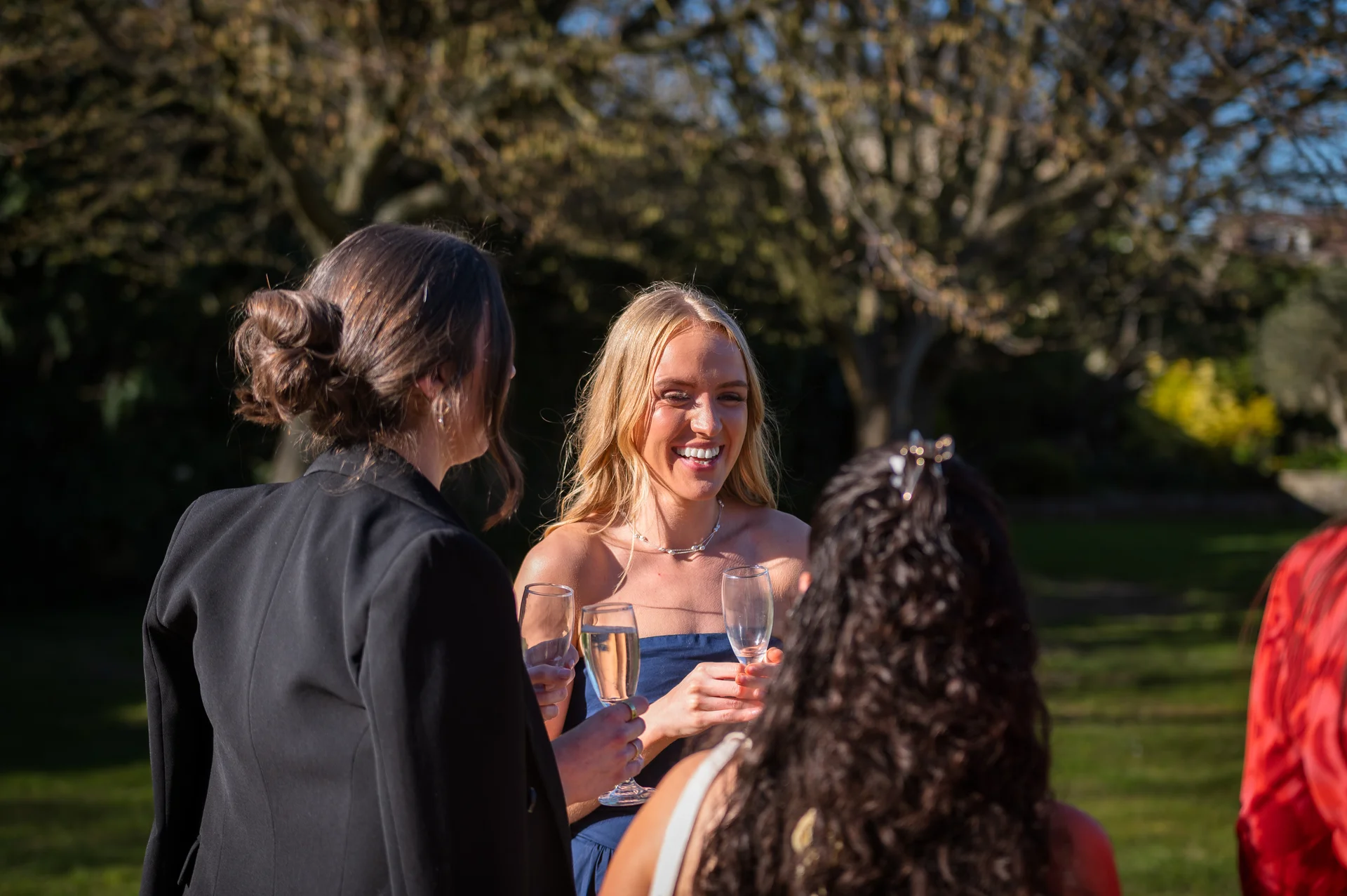 Wedding guests mingle and laugh together outdoors in the sunshine while holding glasses of champagne during the drinks reception.