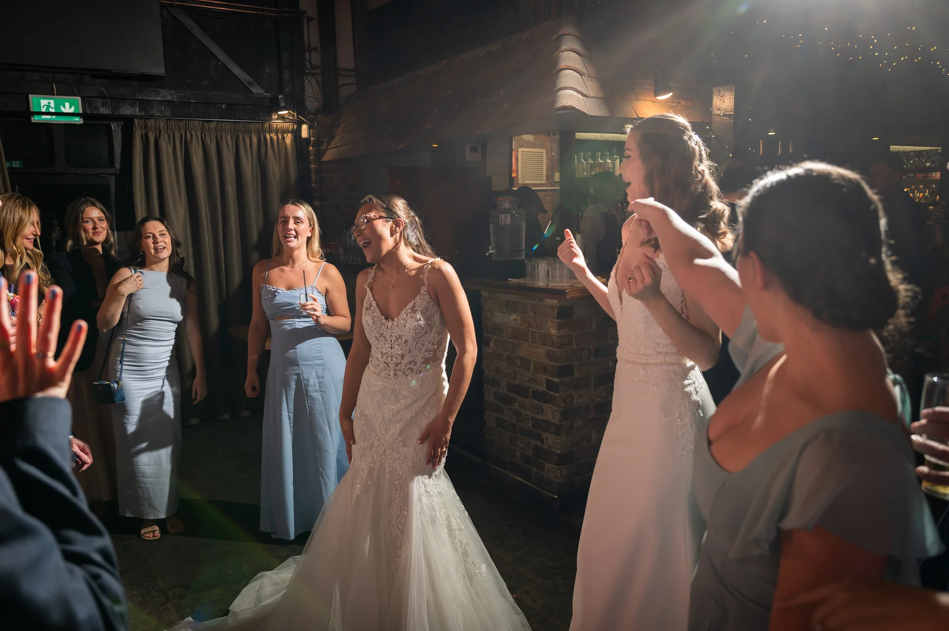 Two brides in white lace wedding dresses laugh and dance joyfully surrounded by guests inside a pub reception venue.