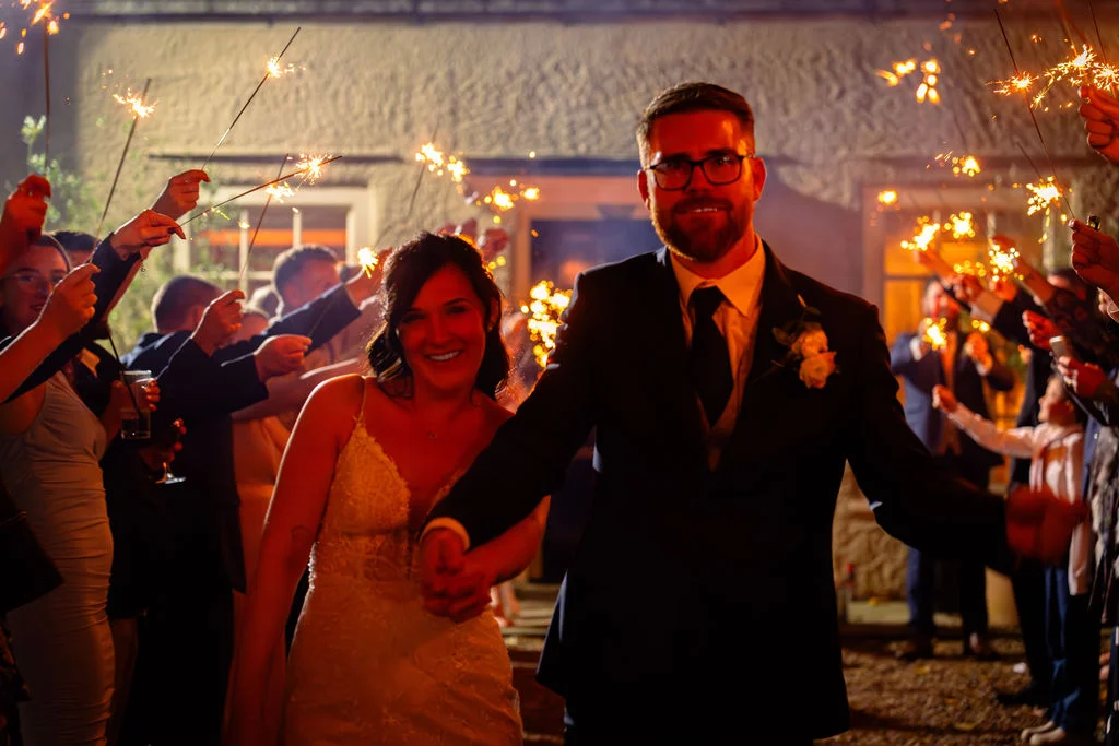 A smiling bride and groom walk hand in hand through a glowing sparkler tunnel held by cheering wedding guests at night.
