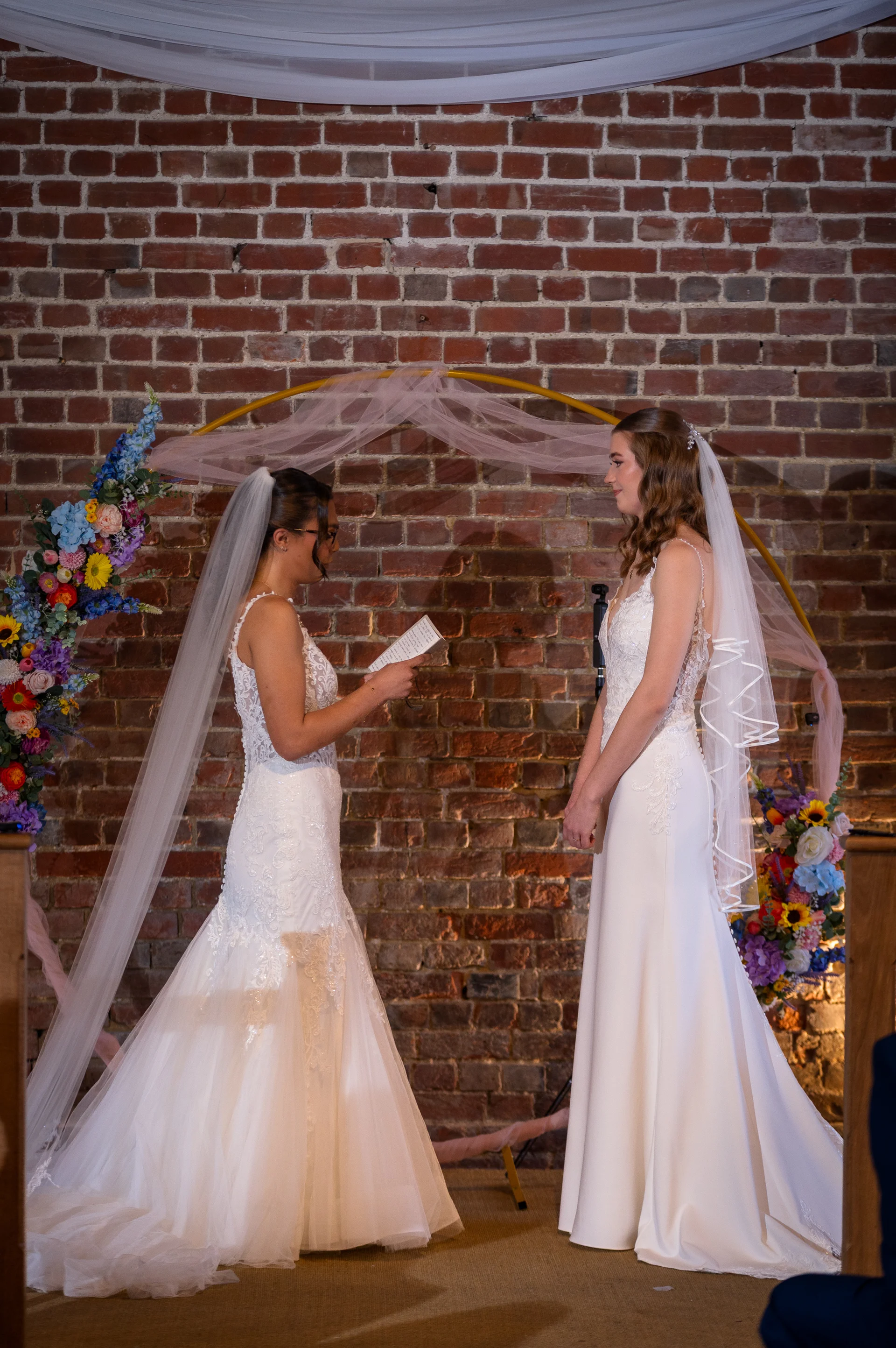 Two brides in white wedding dresses stand facing each other in front of an exposed brick wall and a floral ceremony arch, reading their vows.