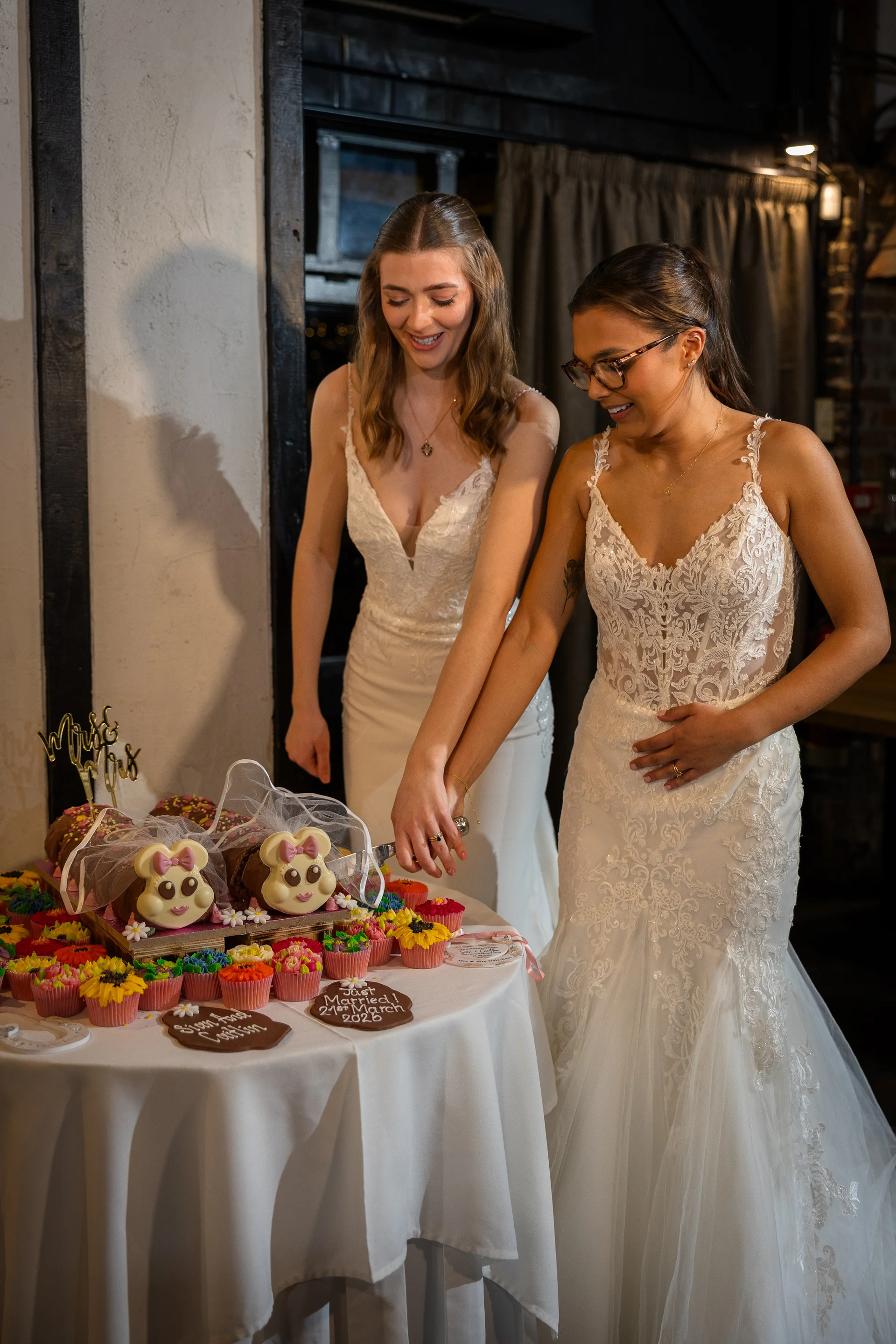 Two smiling brides in lace-accented white wedding gowns stand together cutting a fun themed wedding cake decorated with character faces and cupcakes.