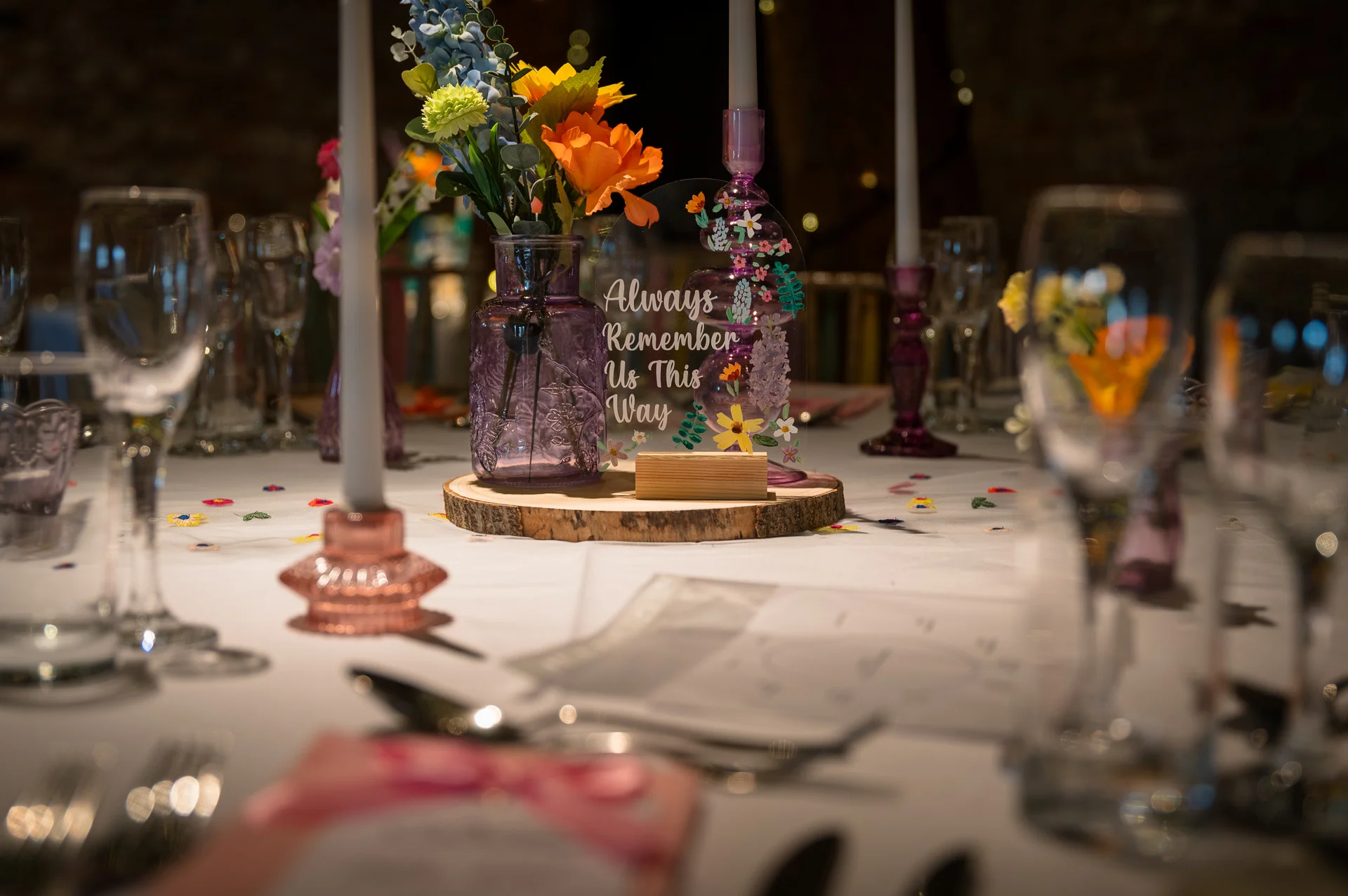 An intimate wedding reception tablescape featuring a wood slice centrepiece with purple vases, colourful wildflowers, taper candles, and an acrylic sign reading always remember us this way.