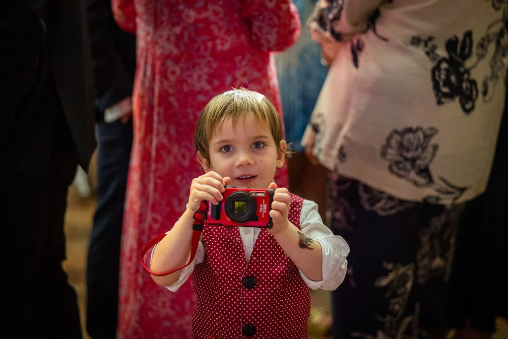 A young page boy wearing a red polka dot waistcoat holds up a small digital camera to take a photo at a wedding reception.