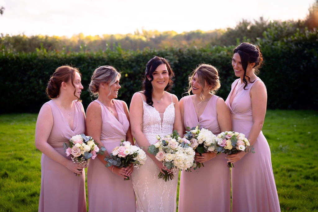 A radiant bride in a lace gown and her four bridesmaids in matching pink dresses stand outdoors on a lush green lawn during golden hour, holding hand-tied floral bouquets and smiling.