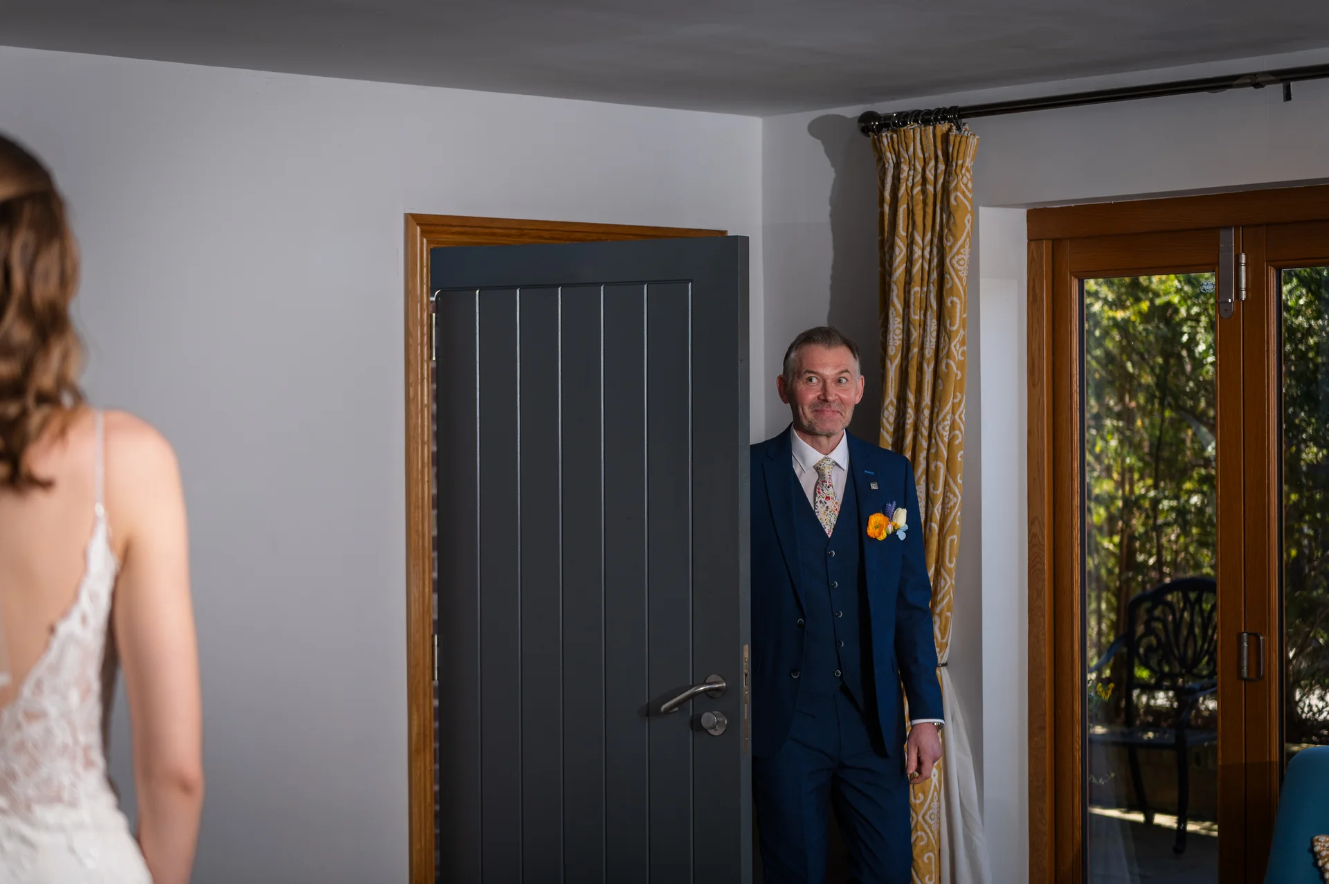 An over-the-shoulder shot captures a father in a blue suit smiling as he sees his daughter in her wedding dress for the first time.