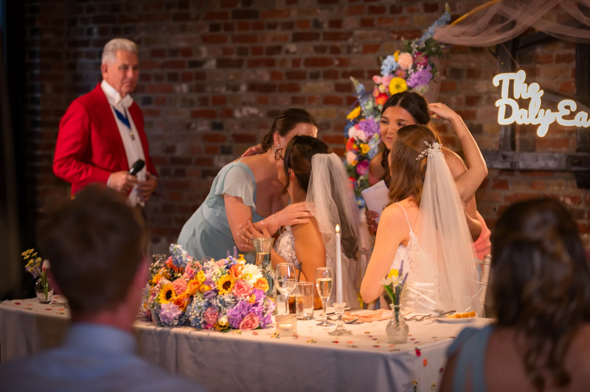 Two brides in lace wedding dresses share an emotional embrace at their top table surrounded by colourful floral arrangements and guests while a toastmaster in a red jacket looks on.