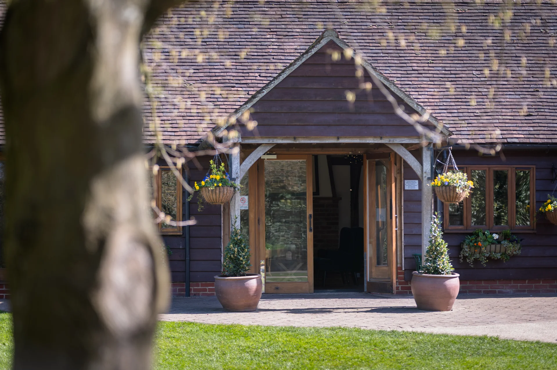 A rustic wooden barn venue with open glass doors, framed by a tree trunk in the foreground and a cobbled courtyard outside.