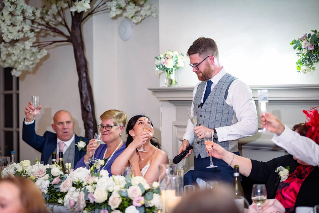 A groom in a tweed waistcoat delivers a speech while seated guests laugh and raise their champagne glasses during the wedding reception.