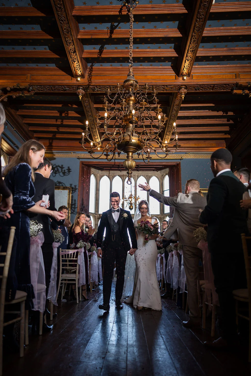 Newlyweds joyfully walk down the aisle hand in hand beneath a grand chandelier in a historic wooden-beamed hall while guests shower them with confetti.