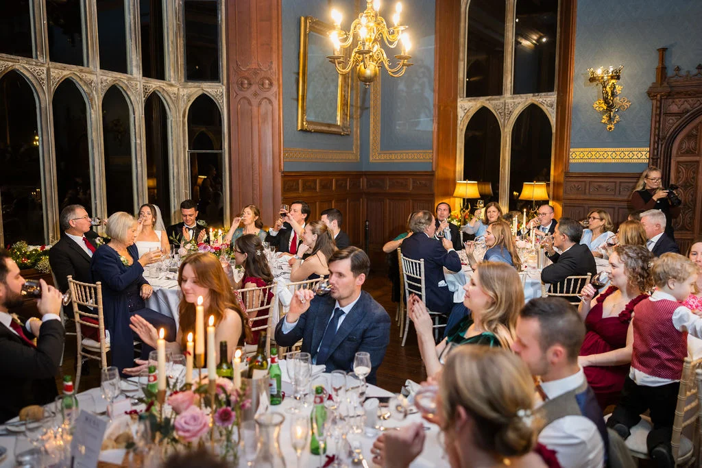 Wedding guests raise their glasses for a toast during a lively wedding breakfast inside an elegant stately home dining room.