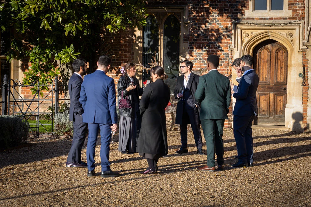 Wedding guests wearing smart suits and coats stand and chat together in a sunlit historic brick courtyard during the drinks reception.