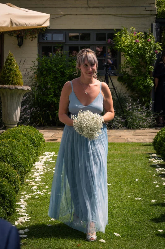 A bridesmaid wearing a dusty blue tulle dress and holding a baby's breath bouquet walks down a garden aisle lined with white petals.
