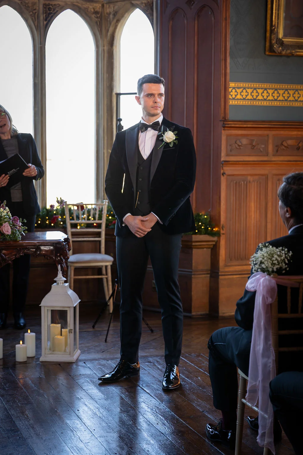 A dashing groom stands at the altar in a velvet tuxedo and black bow tie inside a grand panelled room with tall arched windows.