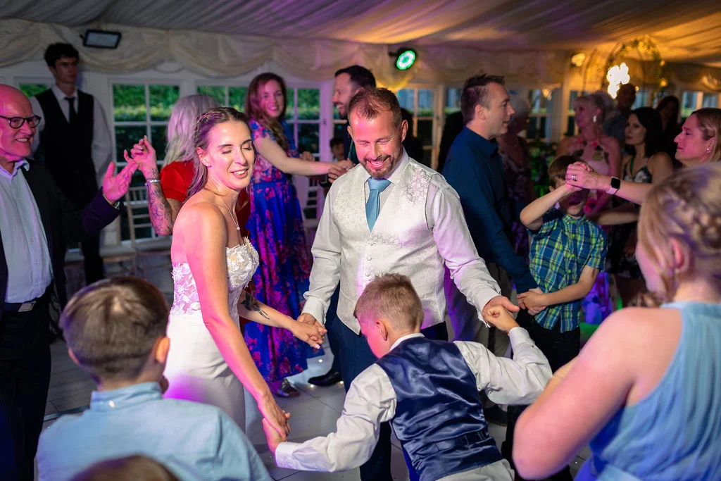 The bride and groom happily dance on the illuminated dance floor with family and young children during a lively evening reception inside a marquee.
