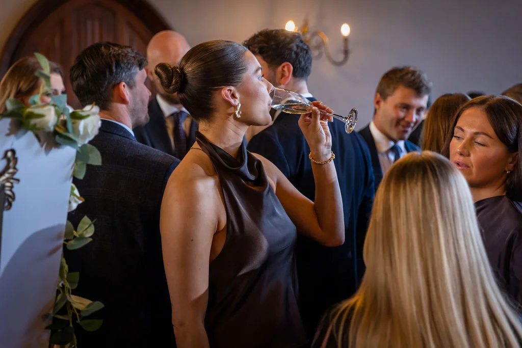 An elegant guest with a sleek updo drinks from a champagne flute while mingling with other guests at a lively wedding drinks reception.