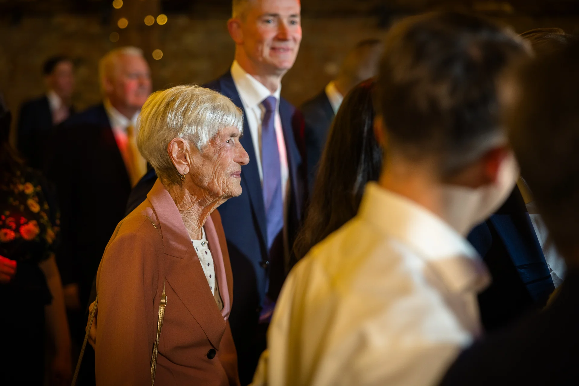 An elderly woman smiling candidly among wedding guests in an indoor venue with warm ambient lighting.