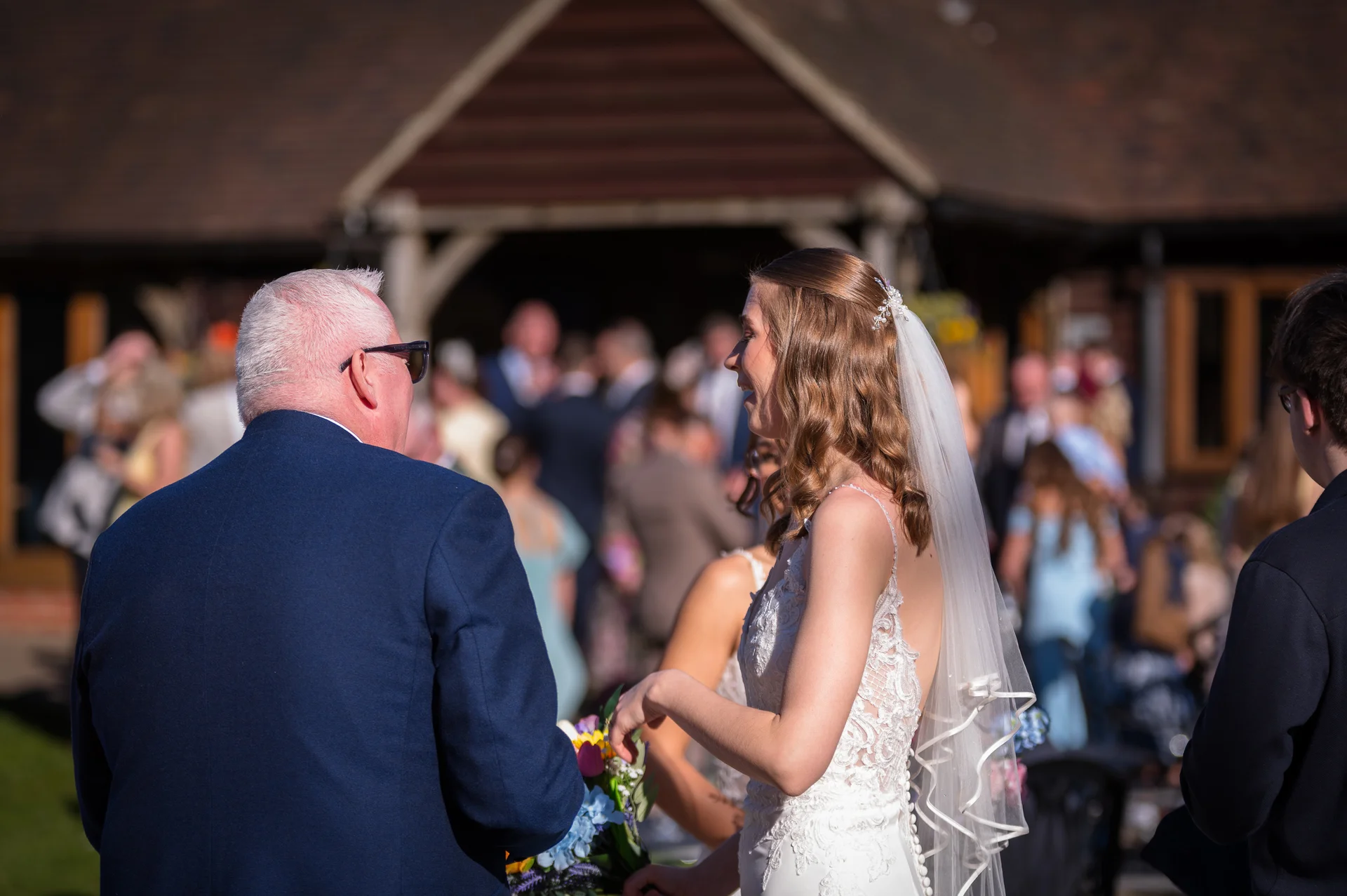 A smiling bride in a lace gown and veil speaks with her father wearing a blue suit outside a rustic barn venue on a sunny day.