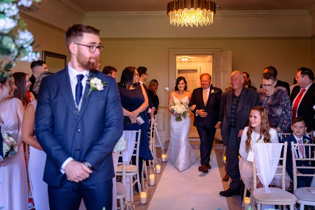 A groom in a navy suit stands at the altar while the bride walks down the aisle accompanied by her father during the indoor wedding ceremony.