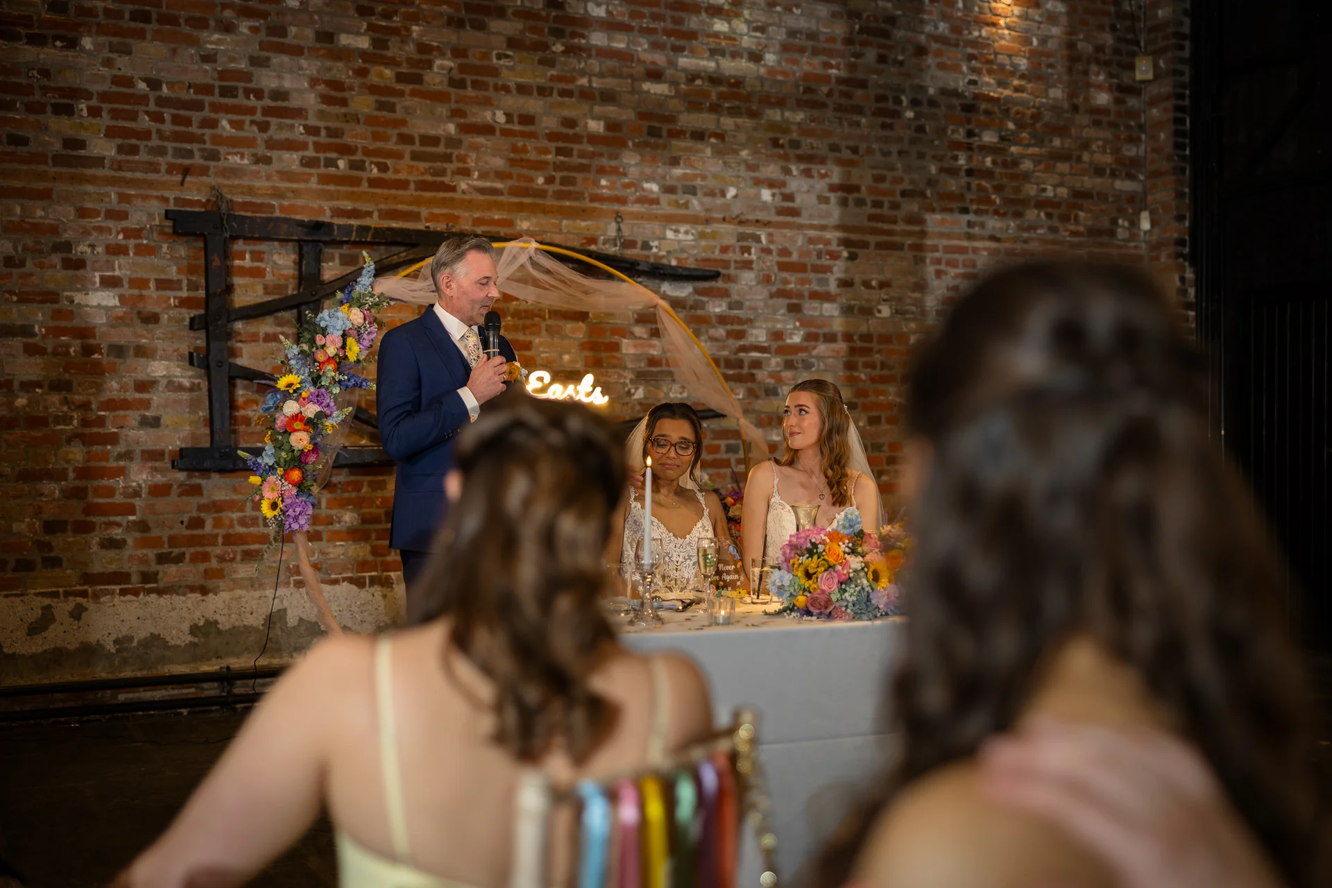 A guest delivers a speech at the top table during a wedding reception, with two brides seated in front of a rustic exposed brick wall and floral arch.