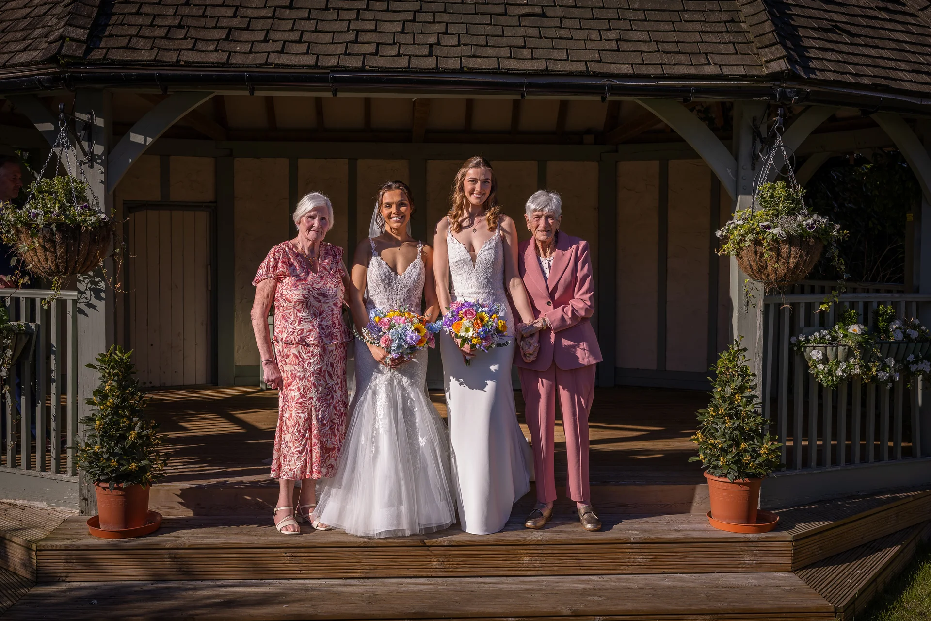 Two brides pose happily with elderly relatives outside a wooden gazebo on their wedding day, holding colourful bouquets.