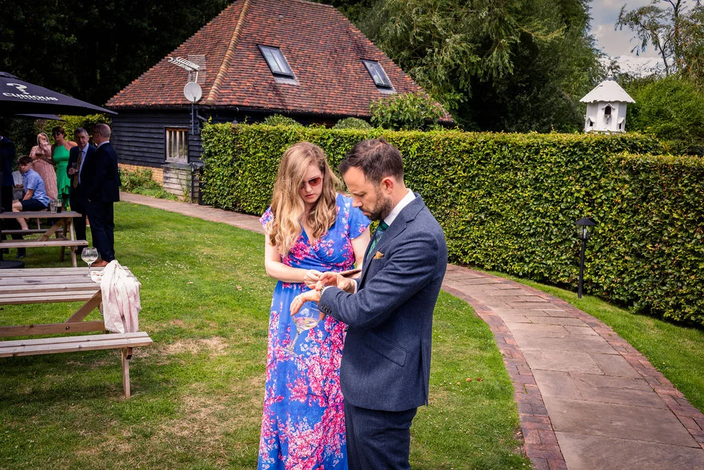 A wedding guest in a grey suit checks his watch next to a woman in a floral blue dress during an outdoor reception near a stone barn.