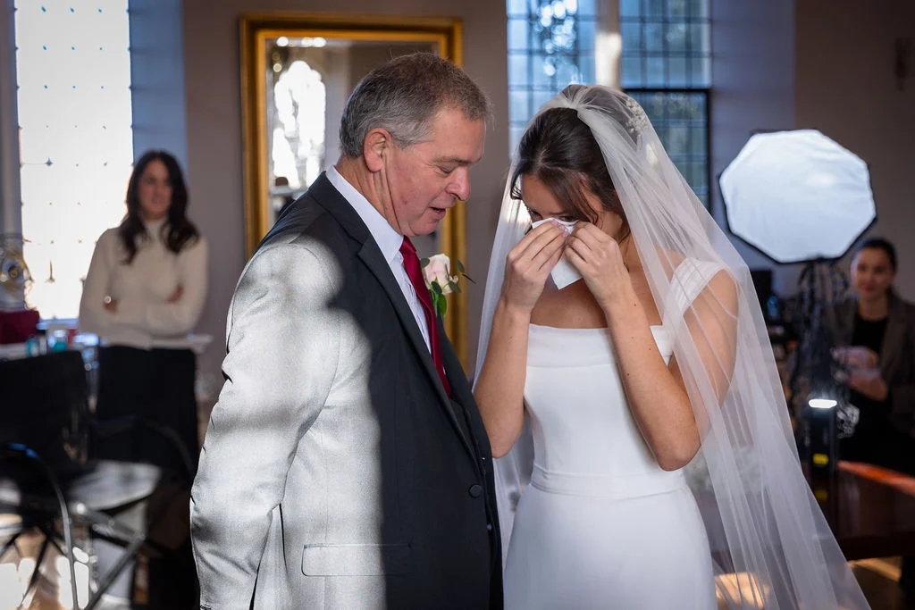 An emotional father of the bride looks on tenderly as the tearful bride gently wipes her eye with a tissue during preparations.