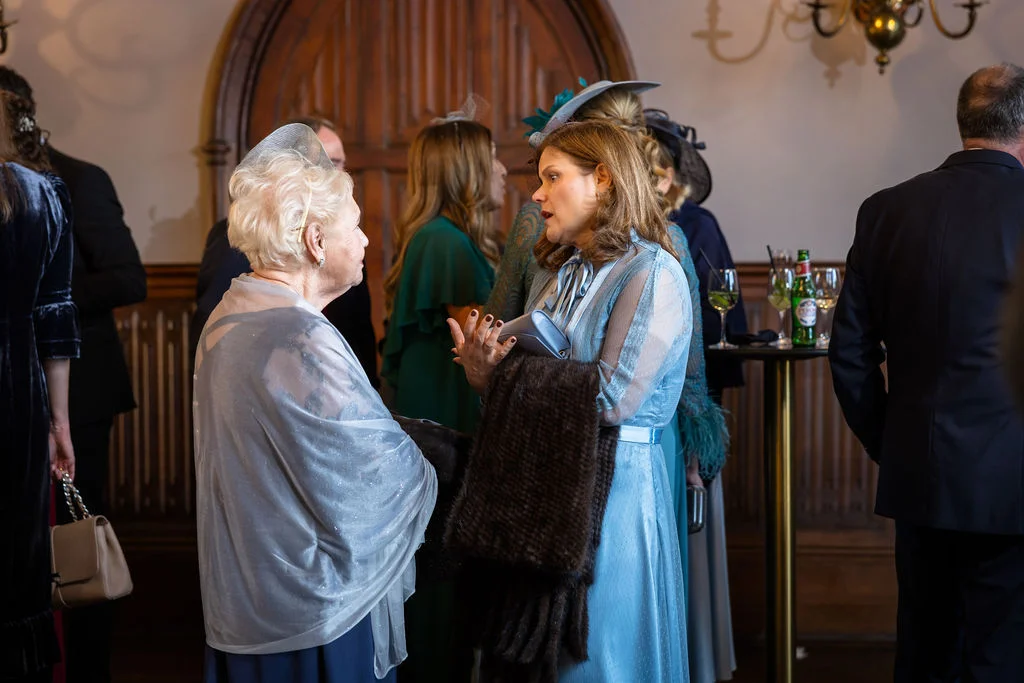 Elegantly dressed wedding guests mingle and converse during the drinks reception in a wood-panelled hall.