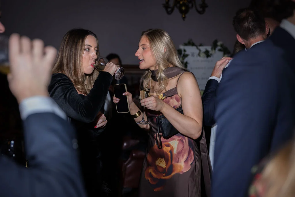 Two female wedding guests chat and drink champagne together at an evening reception surrounded by other attendees.