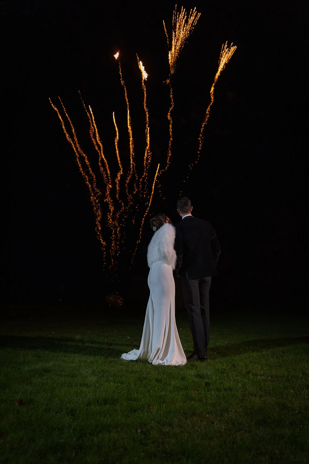A newlywed couple stand on a lawn at night, watching a vibrant golden firework display light up the dark sky.