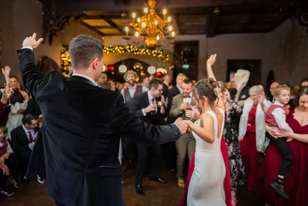 The bride and groom celebrate on a packed dance floor during their evening reception in a grand hall illuminated by a chandelier and festive lights.