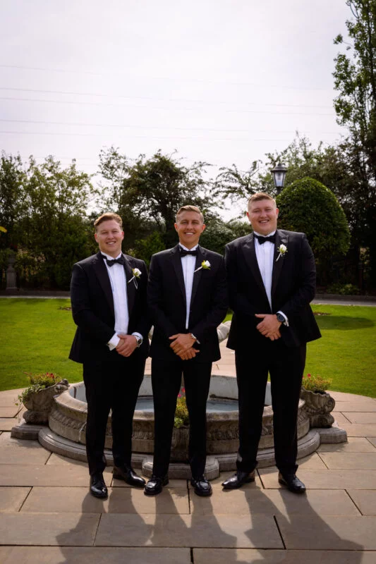 Three smiling groomsmen wearing classic black tuxedos and bow ties pose together outdoors in front of a stone fountain.