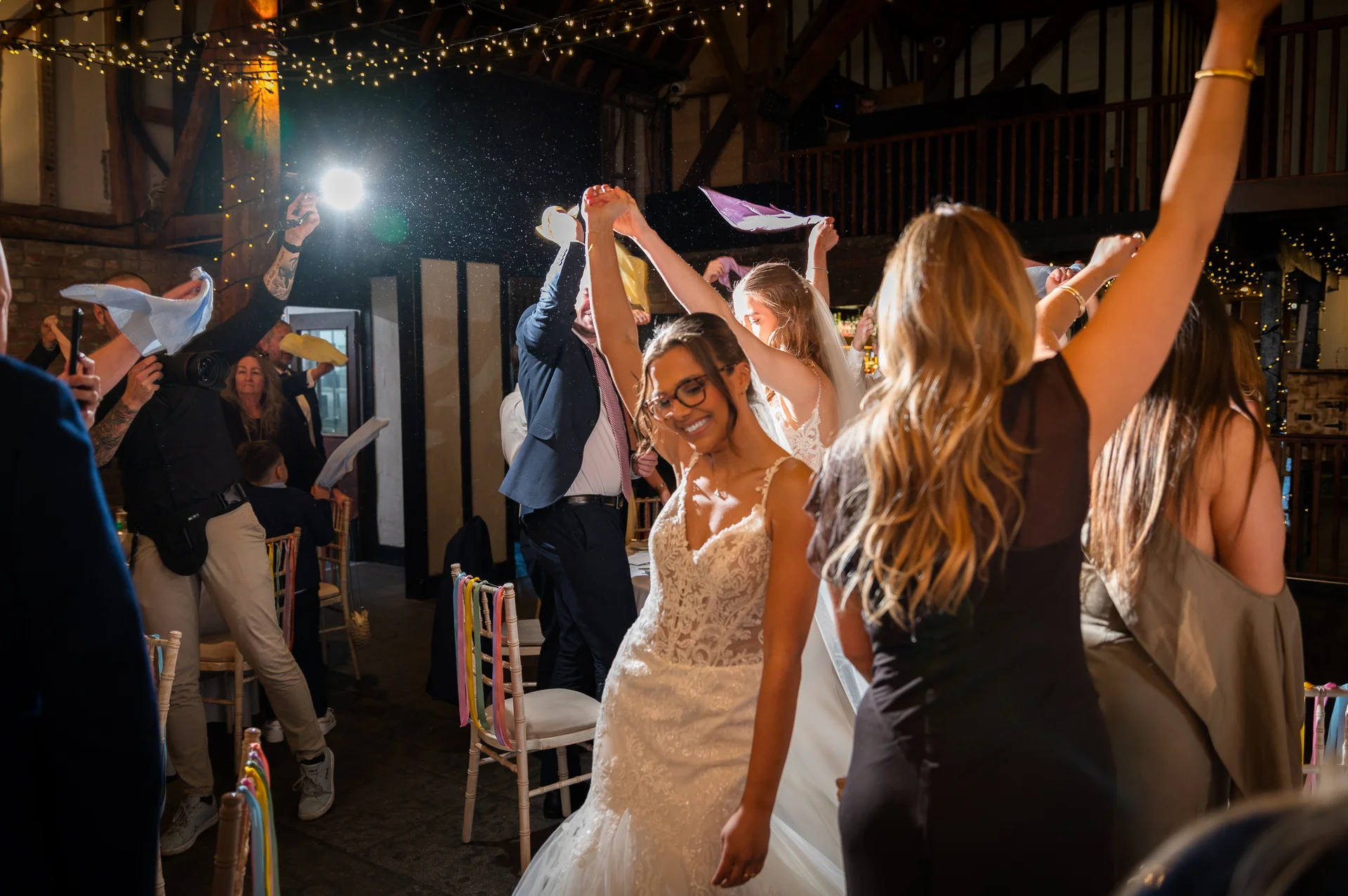 An exuberant bride in a lace wedding gown laughs as guests cheer and wave napkins in the air during an energetic evening reception inside a rustic barn.