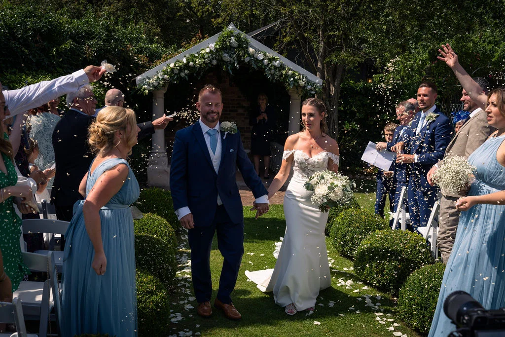 Newlyweds walk hand in hand down the garden aisle while guests enthusiastically shower them with confetti under a sunlit floral arch.