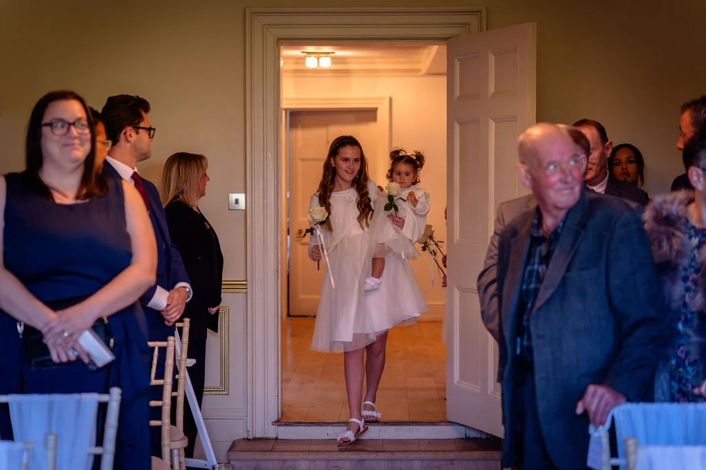 A young flower girl carrying a single white rose walks through an open doorway into a wedding ceremony room while holding a toddler.