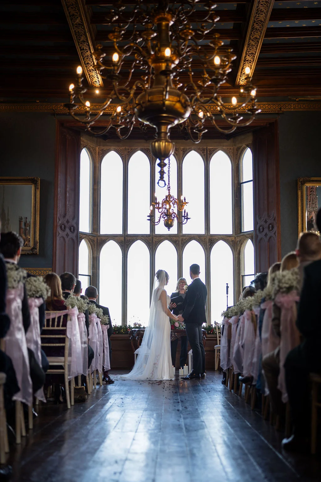 A bride and groom stand exchanging vows in front of a grand arched window inside a magnificent historic hall lit by an ornate chandelier.